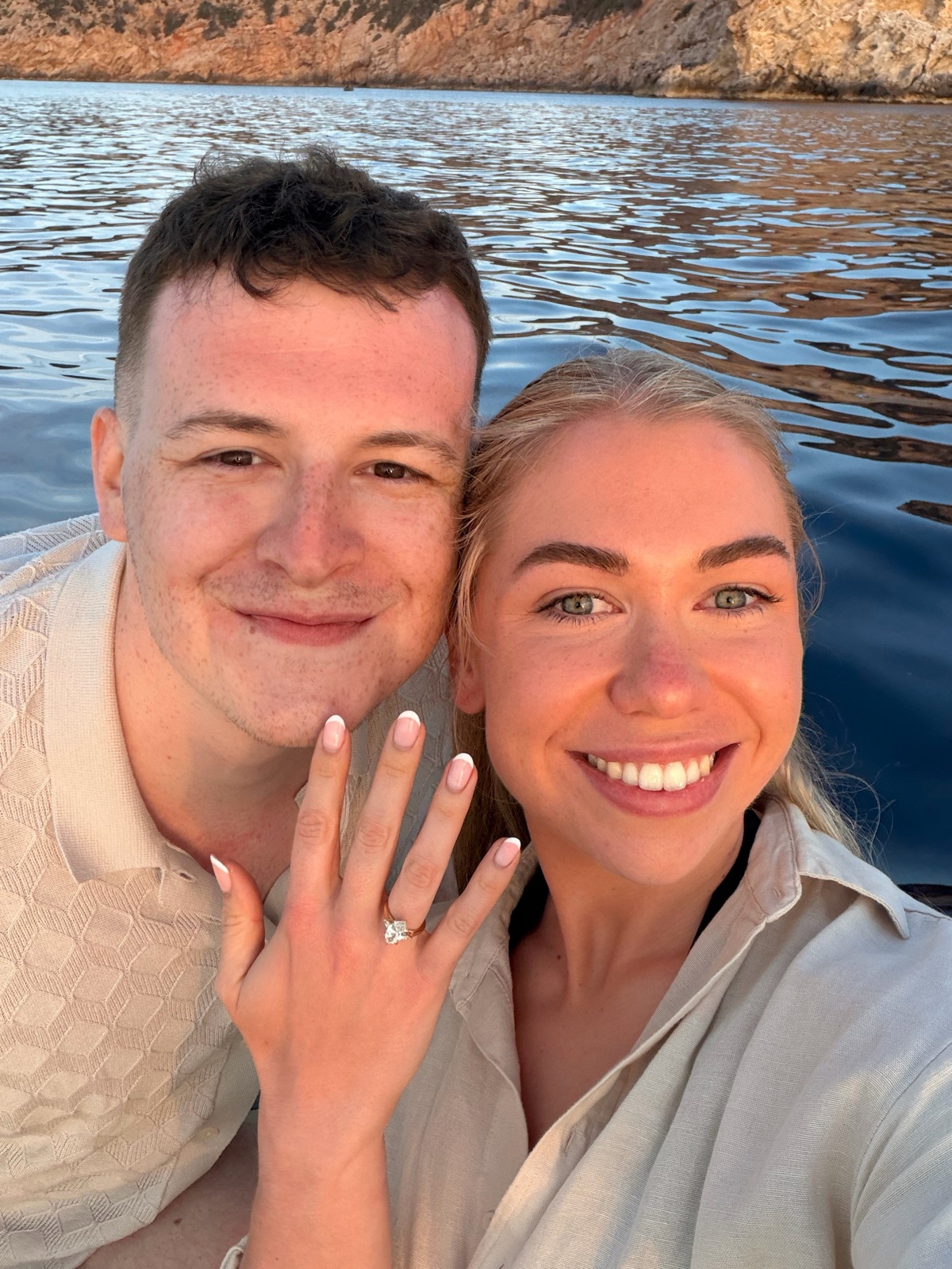 Chloe and Owain smiling by the sea, showing her engagement ring